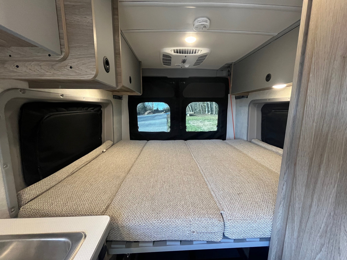 Comfortable RV rental interior with a bed, overhead cabinets, AC unit, and rear windows showcasing outdoor scenery.