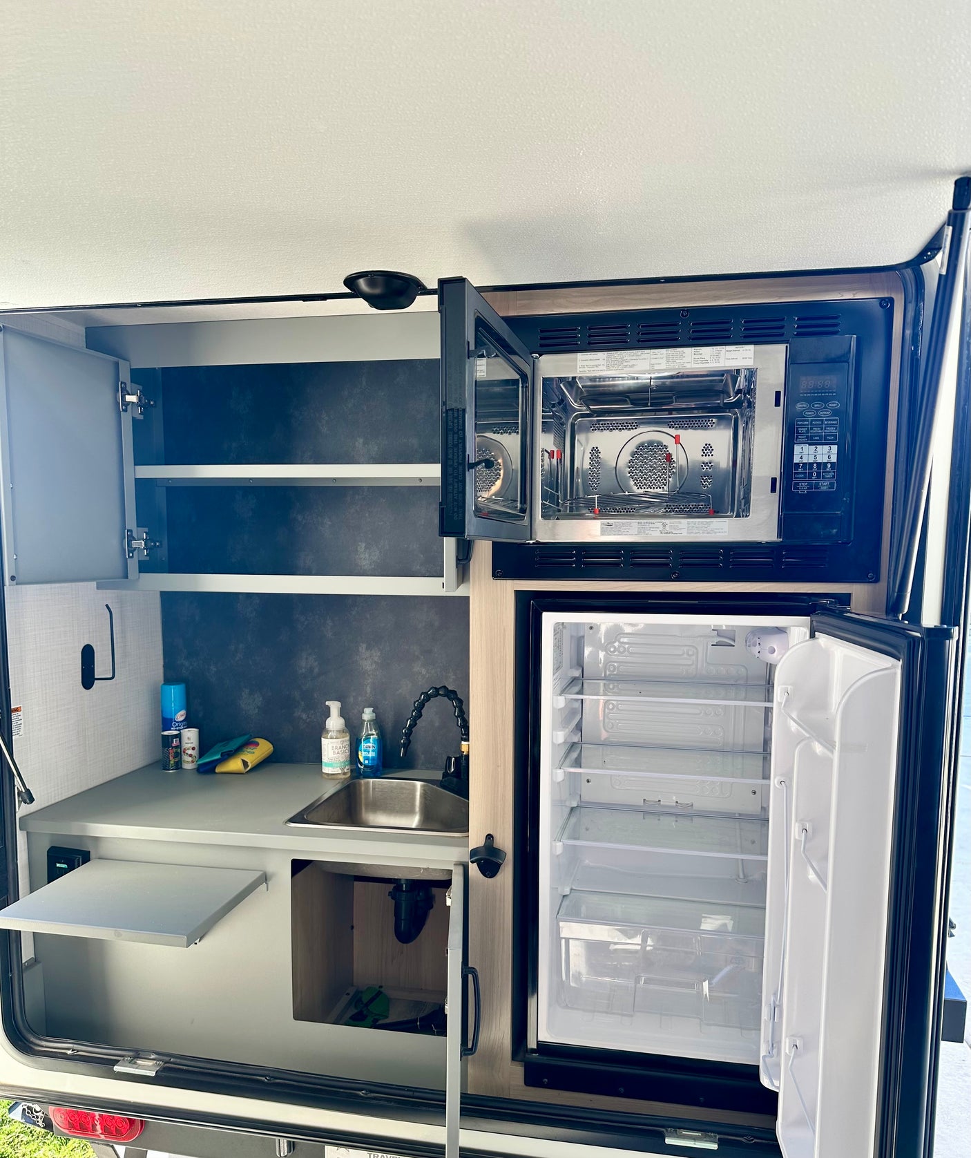 This RV rental's outdoor kitchen features an open microwave, refrigerator, sink, and storage for convenient camping meals.