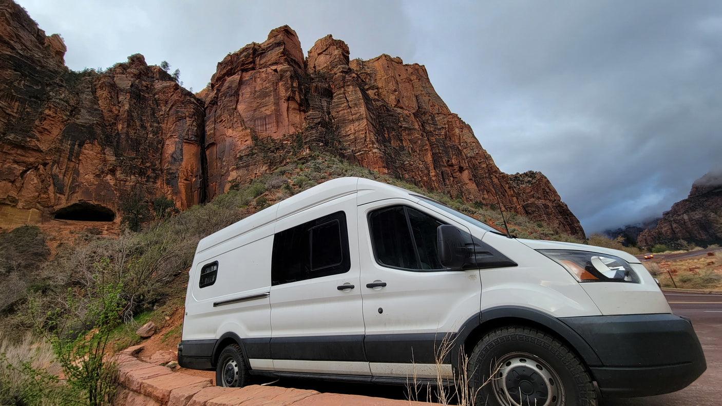 A white **rv rental** camper van parked at Zion National Park's towering red rock cliffs under a cloudy sky, ideal for outdoor adventure.