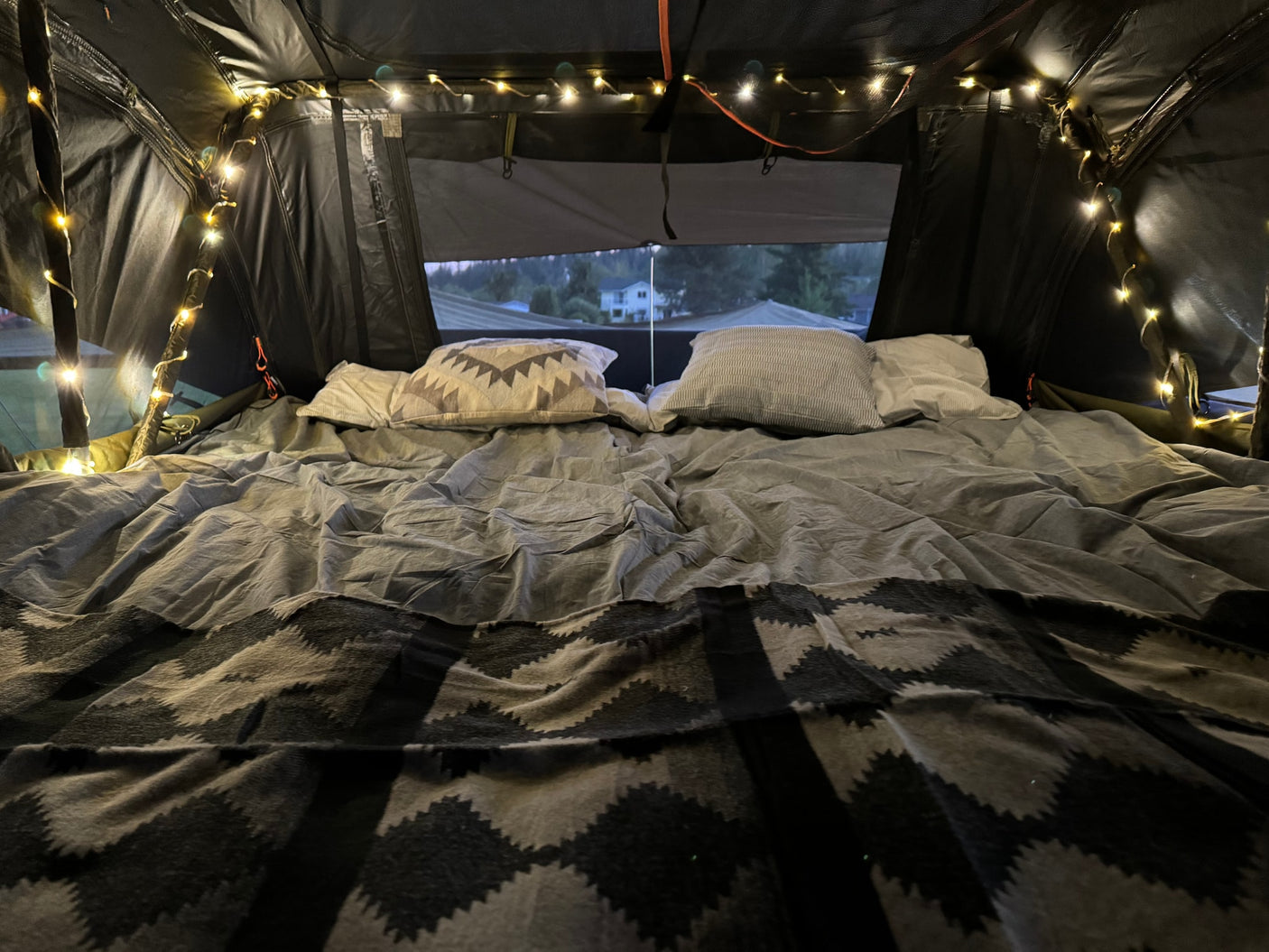 Inside a comfy RV rental: bed with pillows, patterned blanket, and string lights. Views outside from the cozy tent.