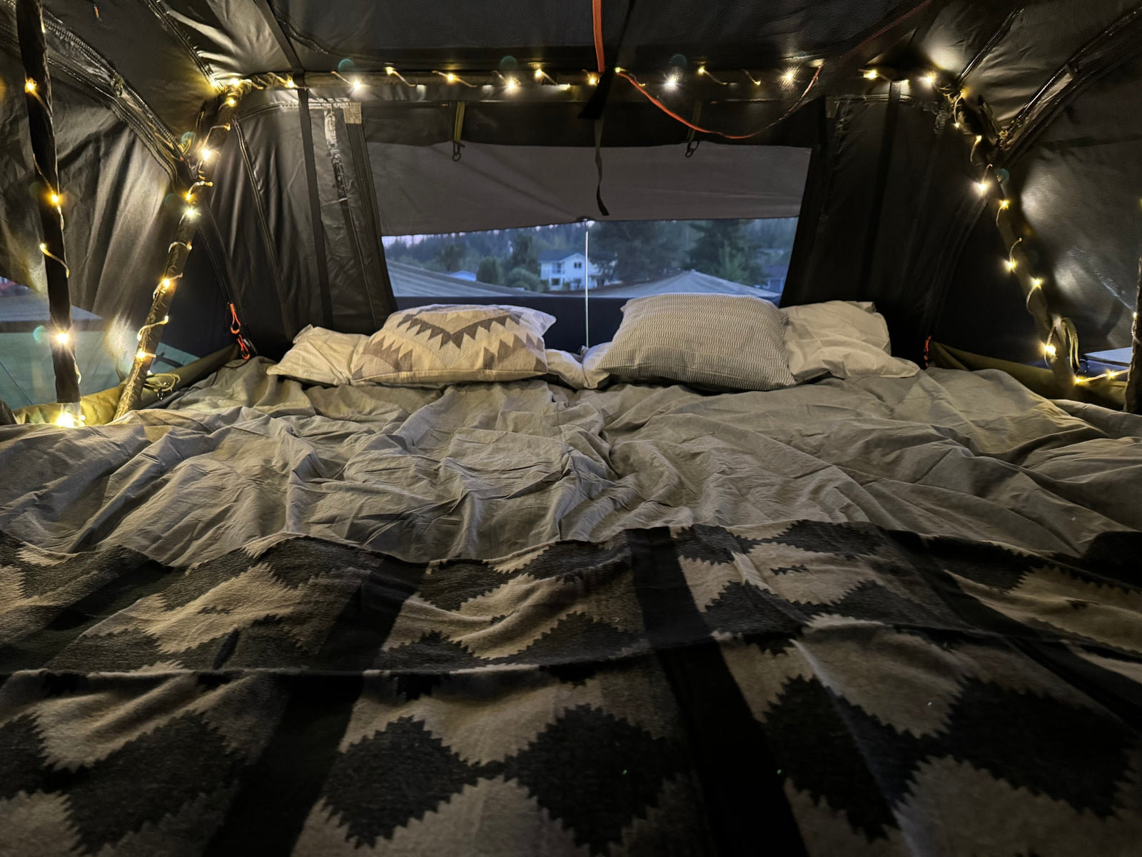 Inside a comfy RV rental: bed with pillows, patterned blanket, and string lights. Views outside from the cozy tent.