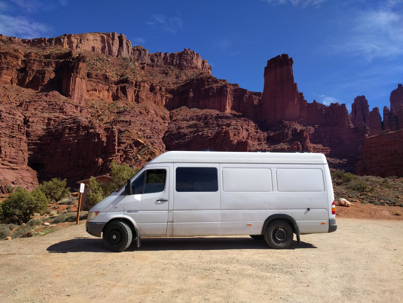 White RV rental van on a dirt road, backed by towering red rock cliffs under a clear blue sky. Ideal for desert road trips.