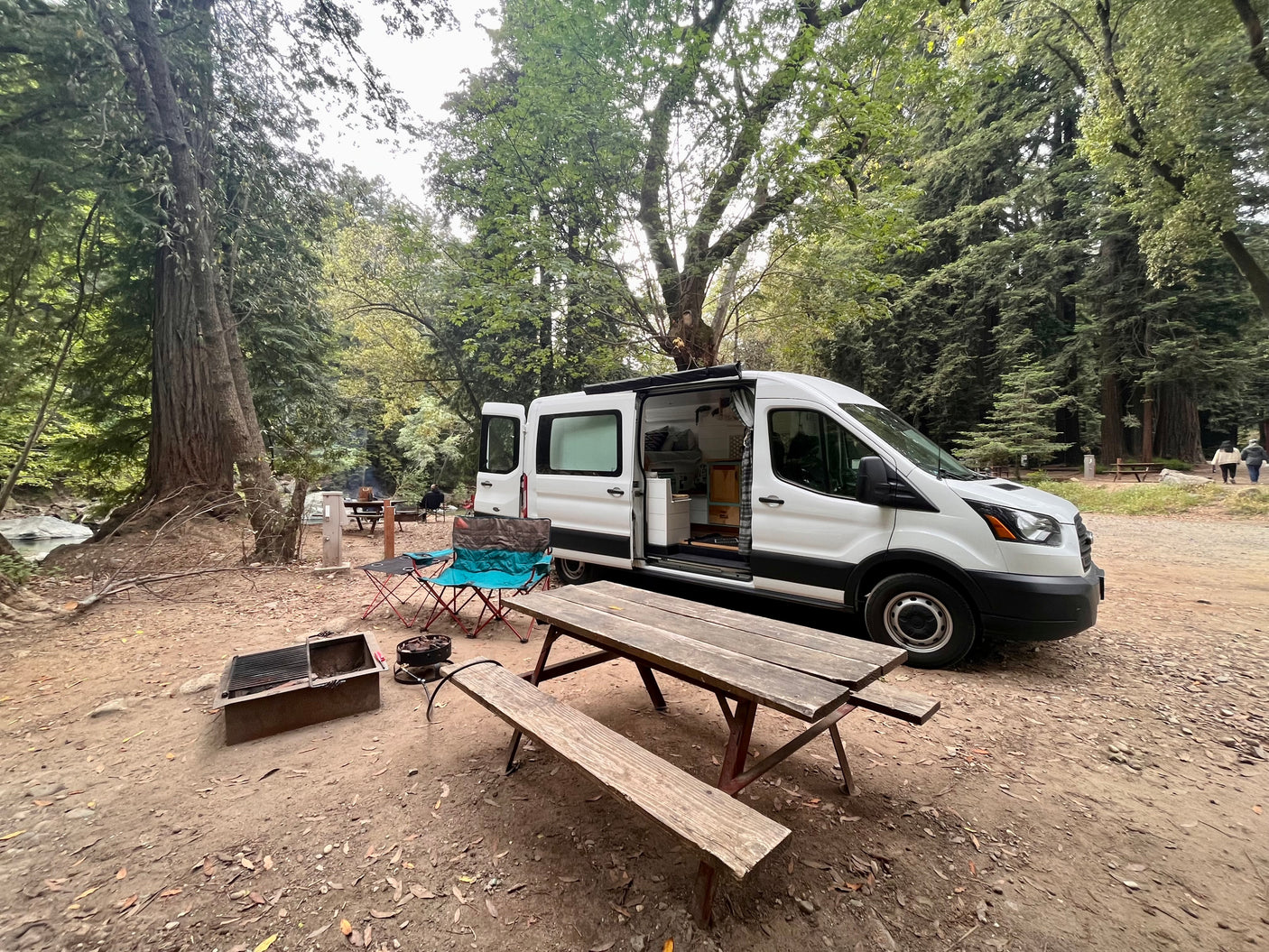 A white camper van, an RV rental, is perfectly set up at a serene forest campsite with a river, picnic table, and fire pit.