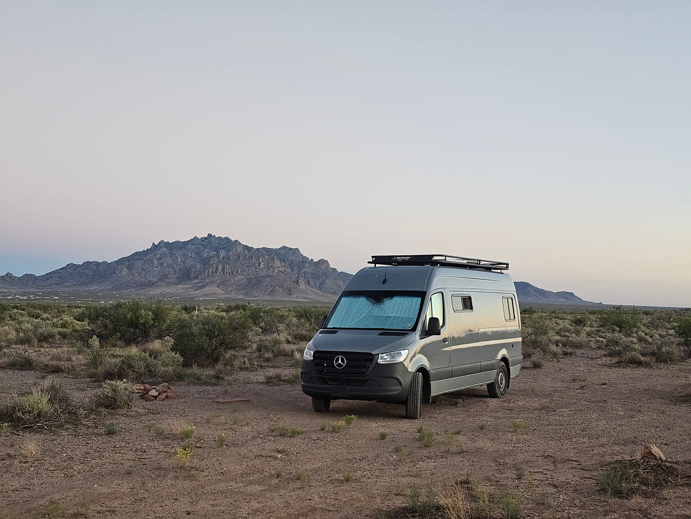 A grey RV rental van with a roof rack is parked in a vast desert landscape, with rugged mountains in the distance under a twilight sky.