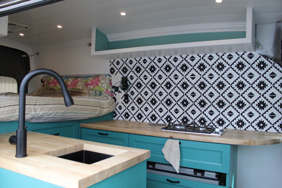 This RV rental interior showcases a cozy custom kitchenette with teal cabinets, wood counters, and a stylish backsplash, next to a raised bed.