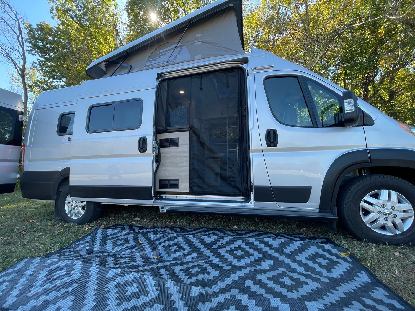 This silver RV rental is seen with its pop-top open, side door screen, and outdoor mat on the grass, ready for camping.