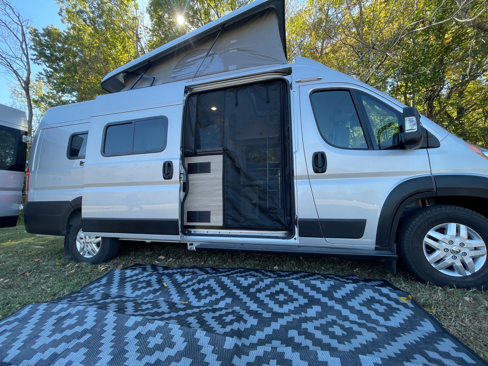 This silver RV rental is seen with its pop-top open, side door screen, and outdoor mat on the grass, ready for camping.