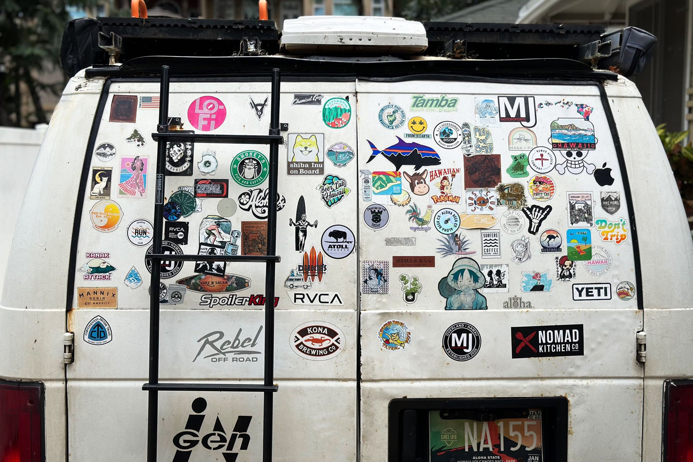 The back of a white RV rental van, adorned with a ladder and countless Hawaii-themed and outdoor adventure stickers.