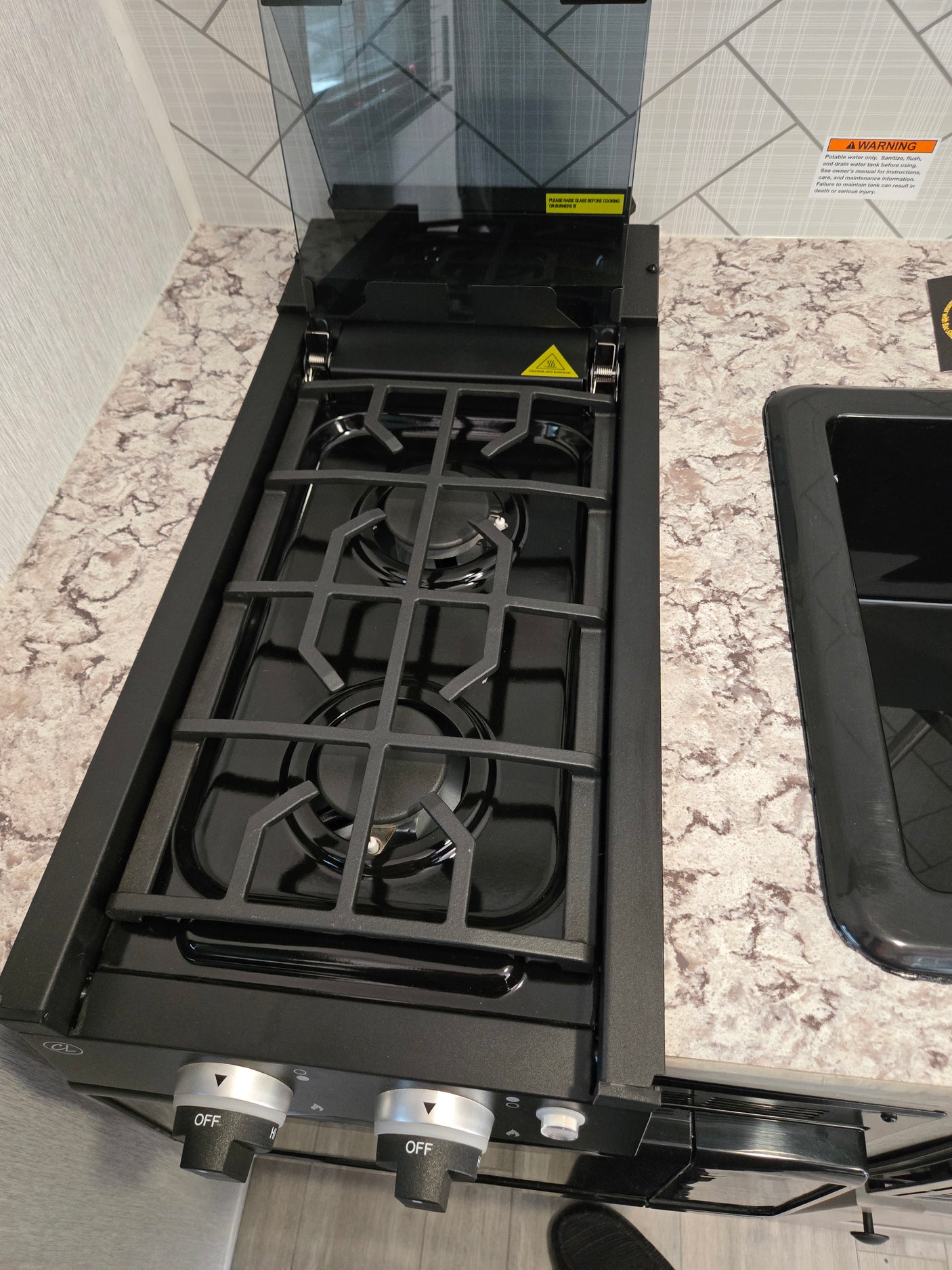 Compact RV rental kitchen features a two-burner gas stovetop with an open glass cover and an adjacent black sink.