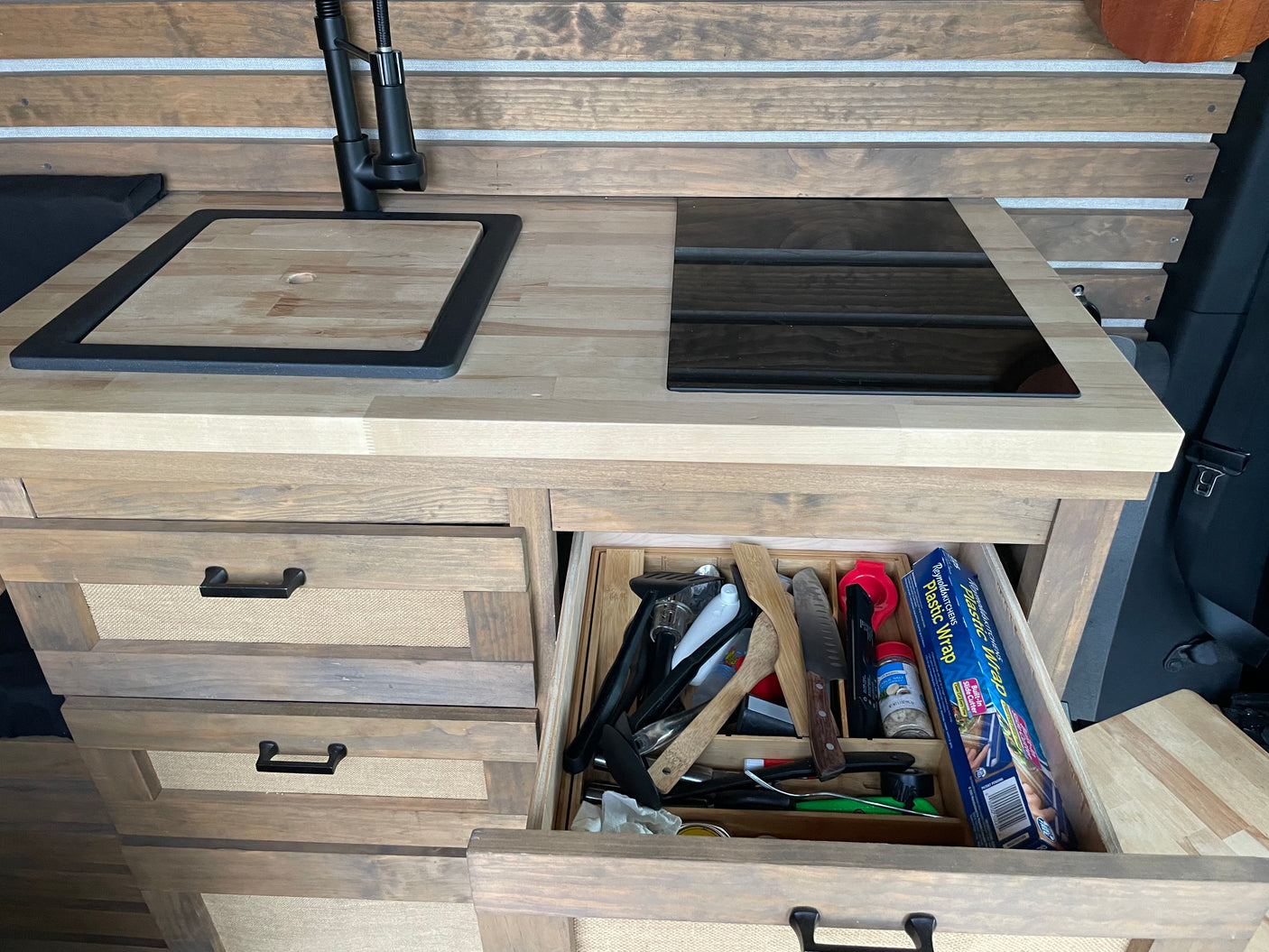 Inside an RV rental: a modern kitchen with wooden counters, sink cover, cooktop, and an open drawer of kitchenware.