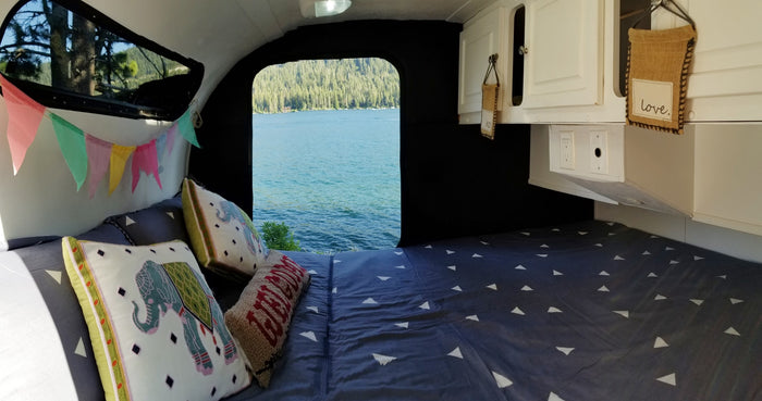 Inside a comfortable RV rental: a cozy bed, colorful bunting, and a serene lake view from the large opening.