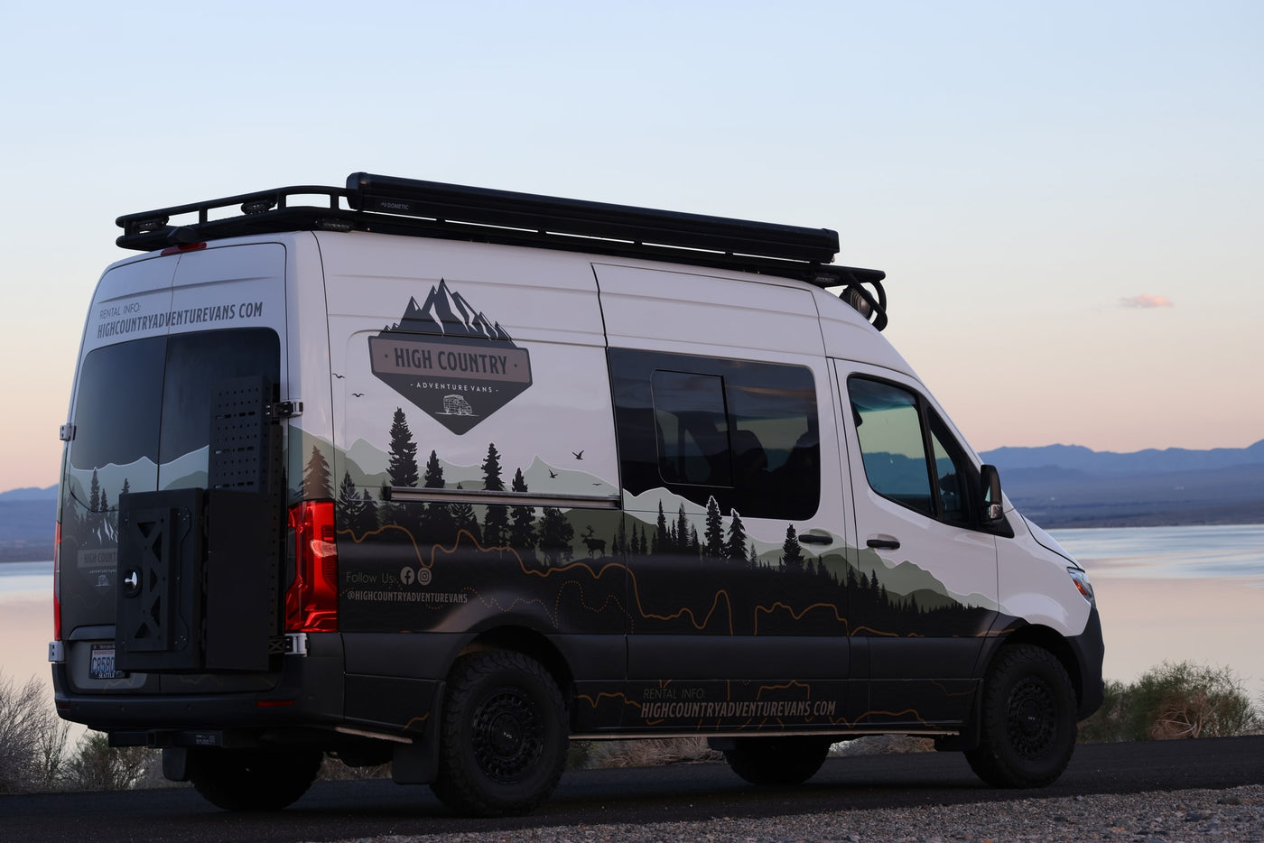 Custom-wrapped adventure rv rental van, with mountain graphics and roof rack, parked by a serene lake at dusk. — photo 1