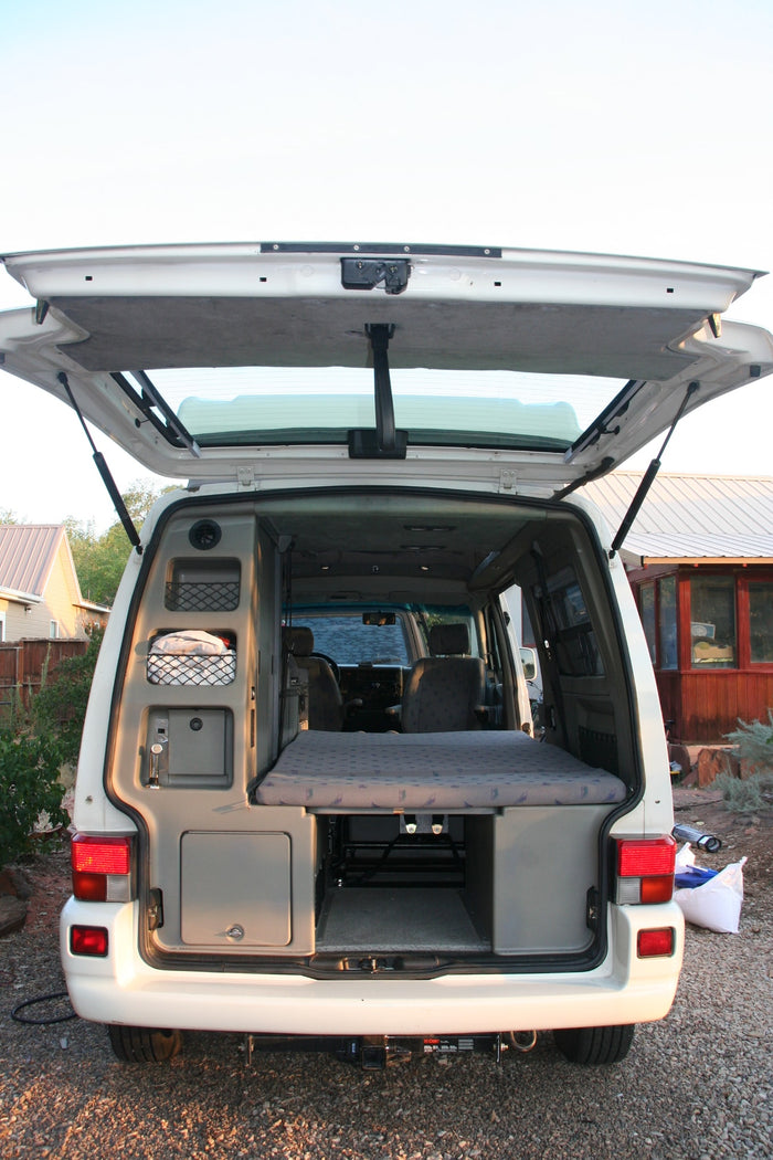 Open rear of a white camper van, revealing its interior setup with folded bed and storage. Ideal for an RV rental.