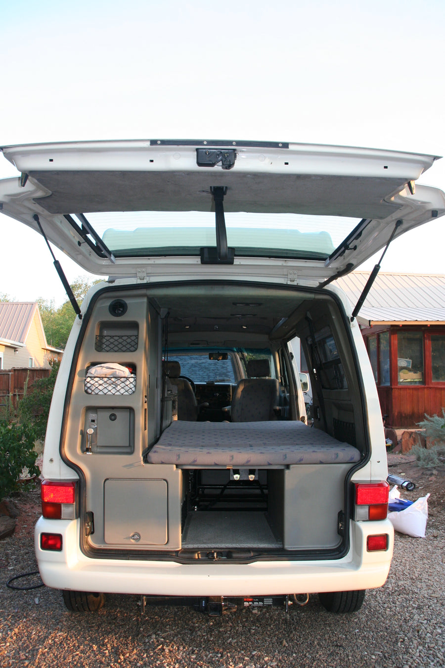 Open rear of a white camper van, revealing its interior setup with folded bed and storage. Ideal for an RV rental.