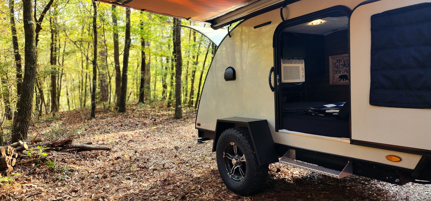 Enjoy nature! This cozy teardrop RV rental, parked in a vibrant forest, welcomes you with an open door to a bed and AC.