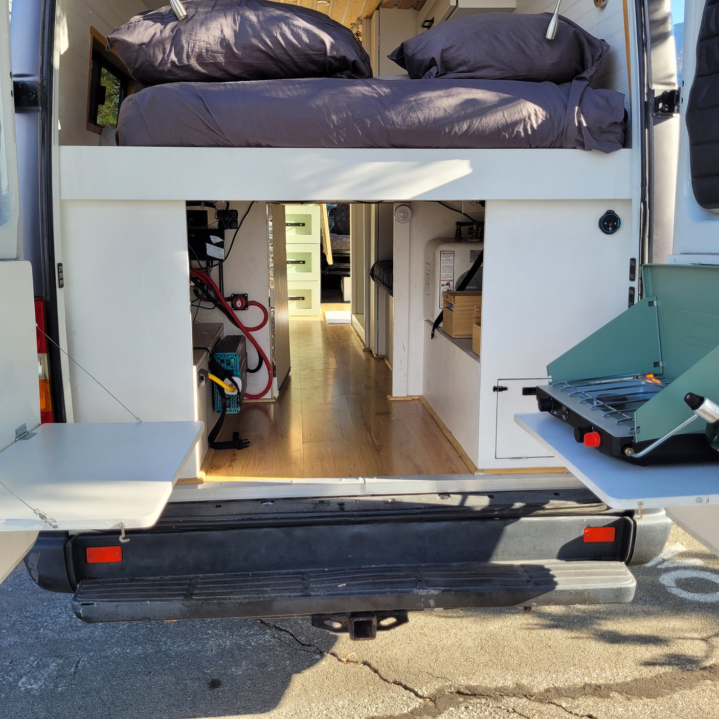 Inside an RV rental: a raised bed, under-bed storage with power and water systems, plus a rear outdoor kitchen with a portable stove.