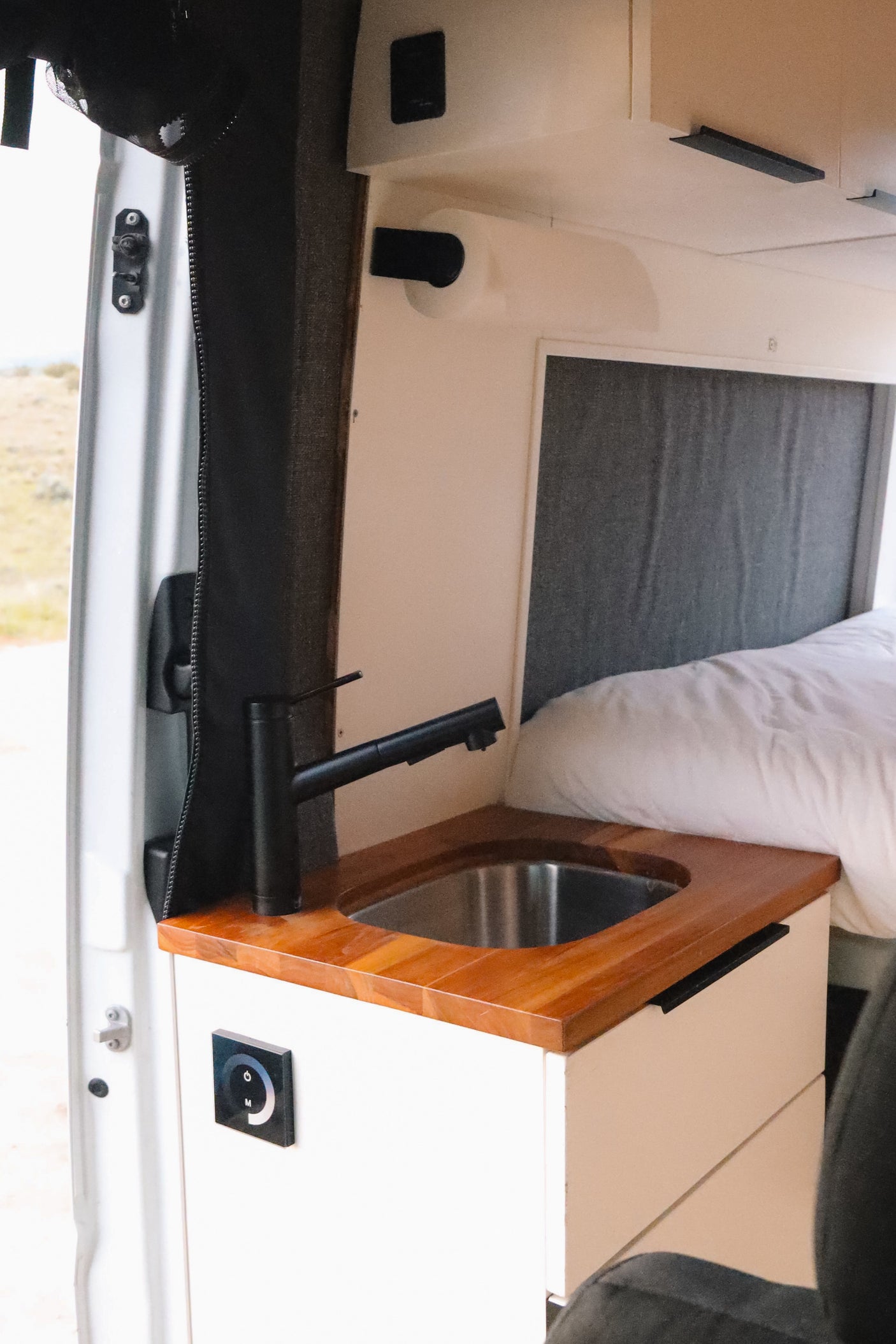 Inside this rv rental, a modern sink, wooden counter, and functional control panel are next to a cozy bed.