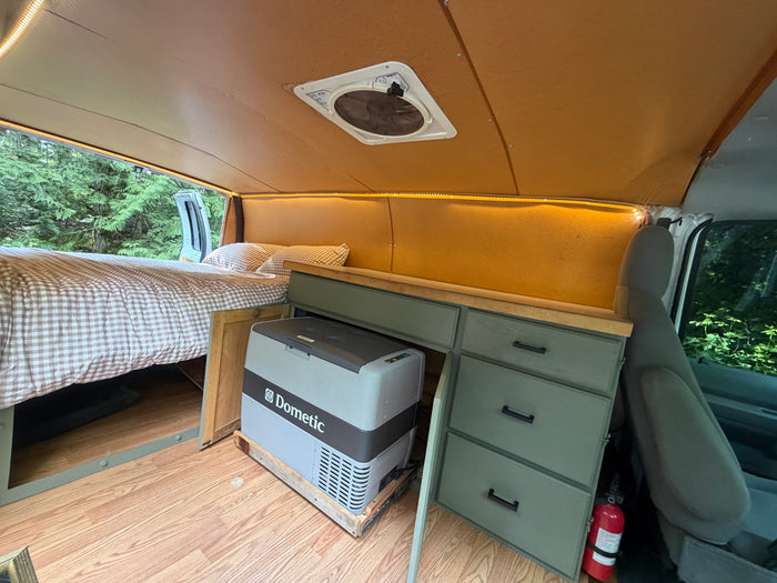 Inside a custom rv rental: a cozy bed, Dometic fridge on a slide, and sage green cabinets offer comfortable living.