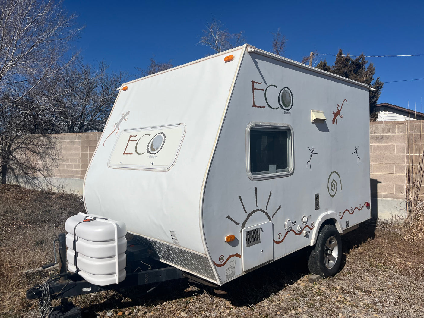 White Eco Skamper RV rental: travel trailer with lizard & sun decals, parked outdoors under blue sky.