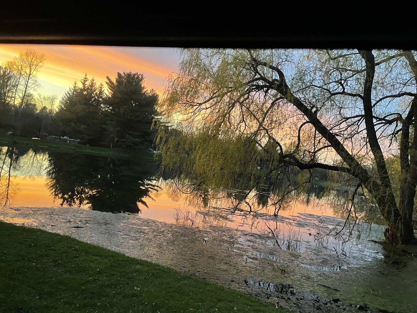 Golden sunset over a tranquil lake, trees reflecting softly, with an RV rental parked on the distant shore.