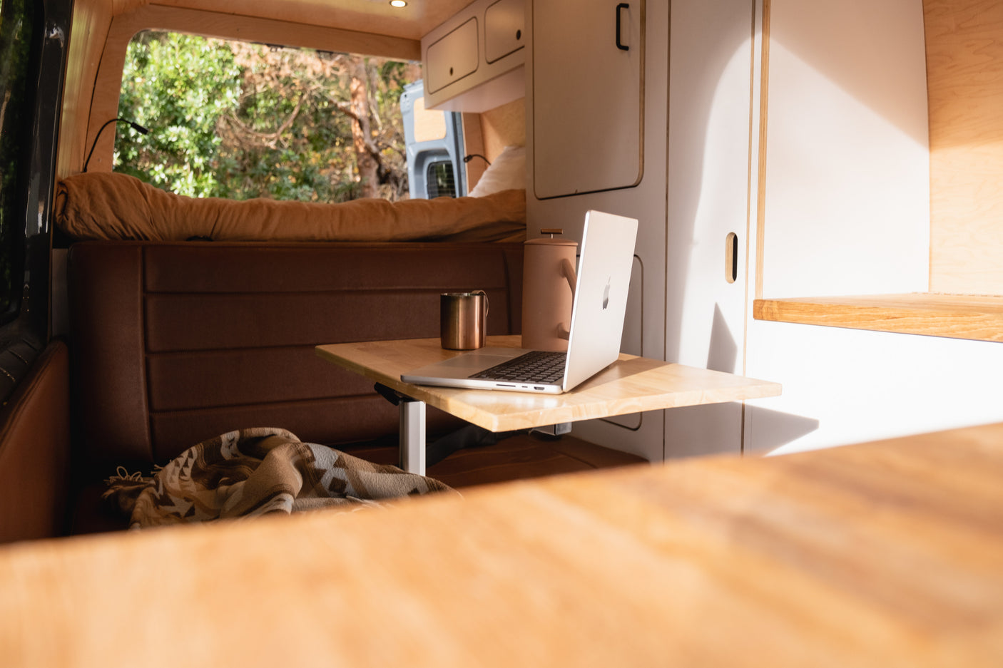 Cozy RV rental interior shows a camper van workspace with a laptop, coffee, and a comfy bench overlooking nature.