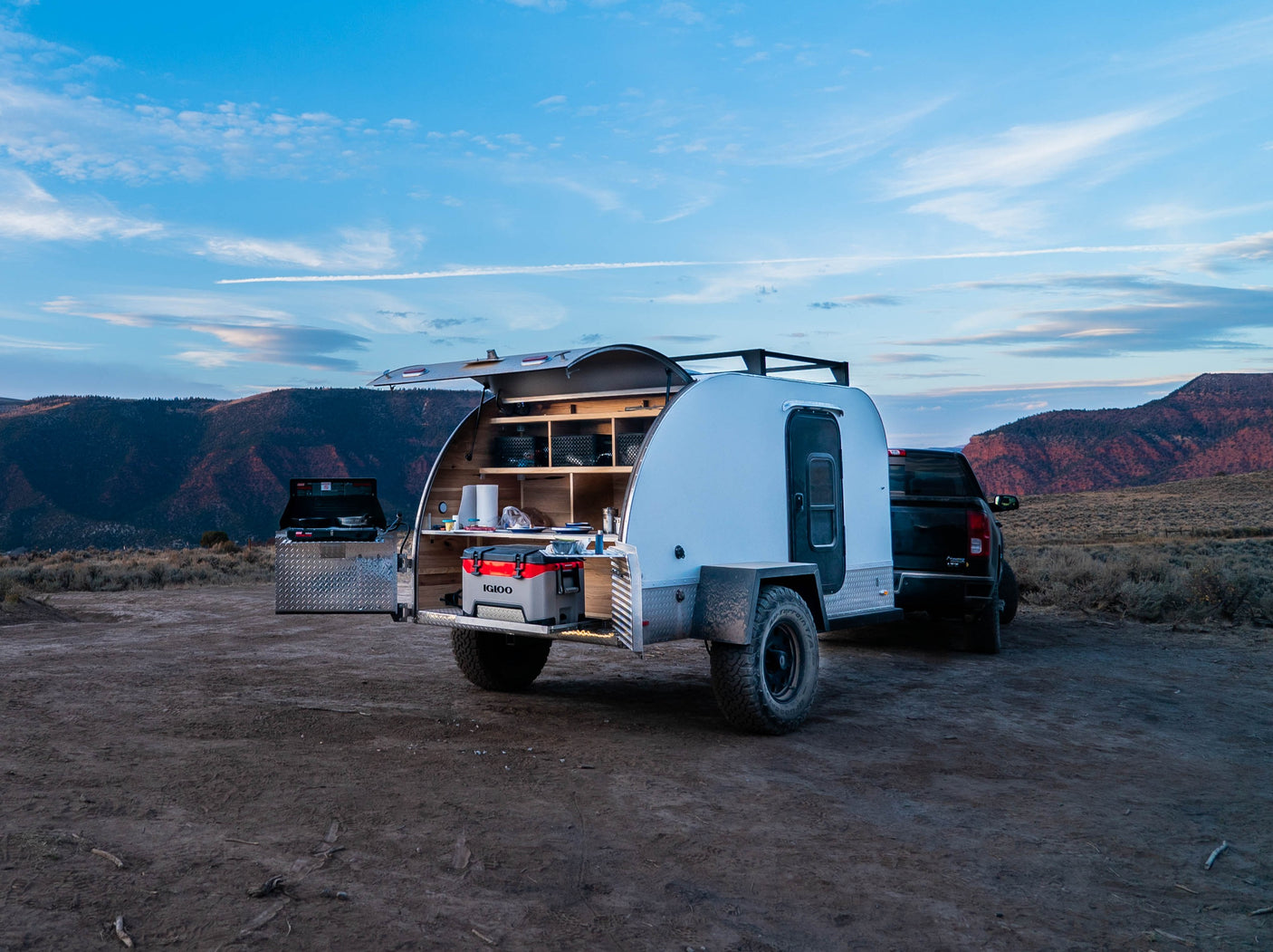 Outdoor rv rental adventure: A white teardrop camper with an open rear kitchen, including an Igloo cooler, hitched to a truck at dusk.