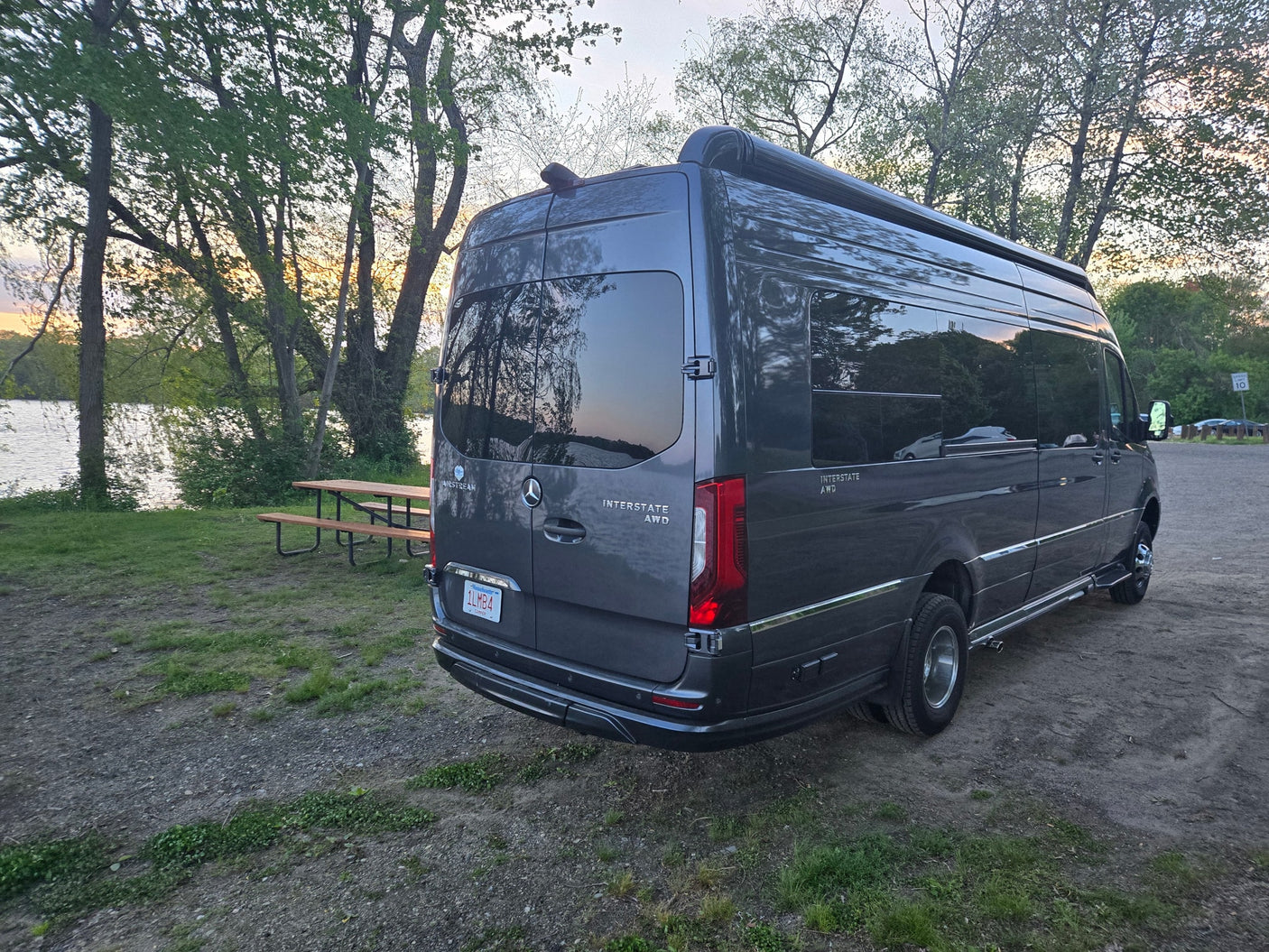 Experience nature with an RV rental! A gray Airstream Interstate AWD campervan parked by a lake and picnic table at sunset.