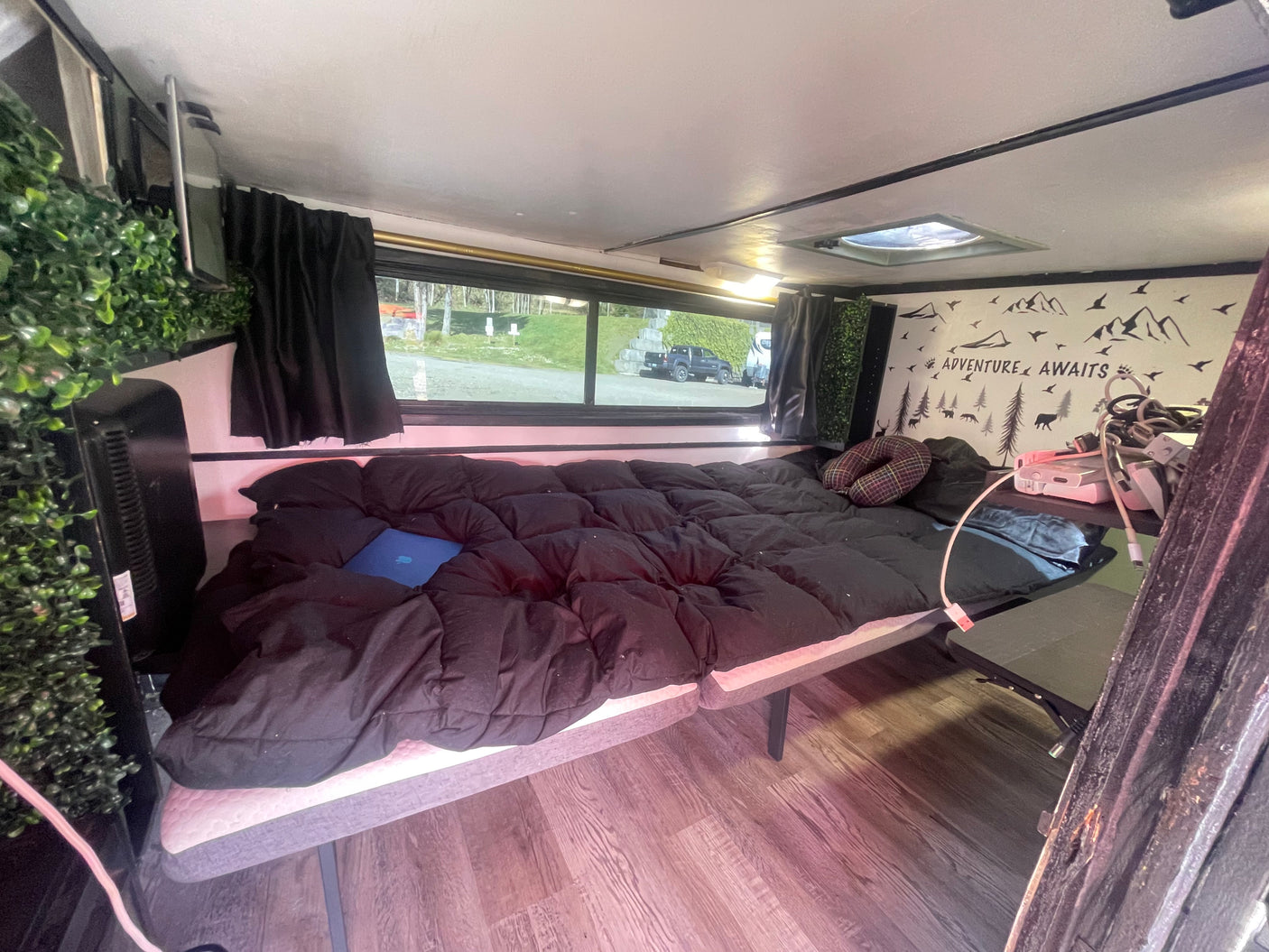 Inside an RV rental: a compact living space featuring a comfy bed, window view, and "Adventure Awaits" wall art.