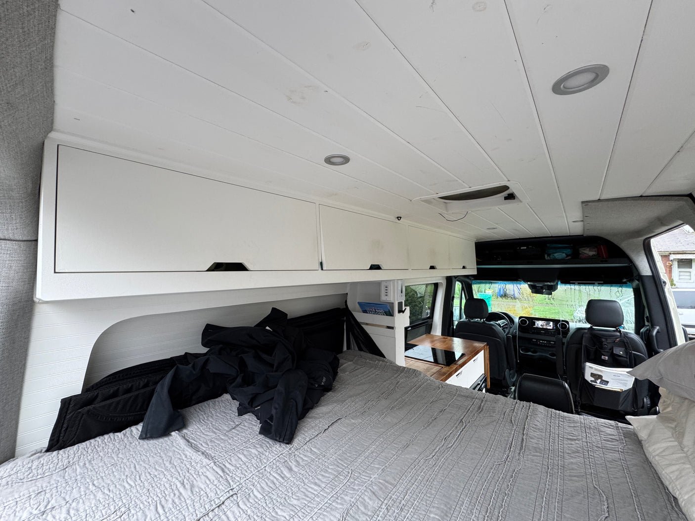 View from the back of an RV rental, showing a grey bed, white overhead cabinets, and a compact kitchen counter, leading to the front cab.