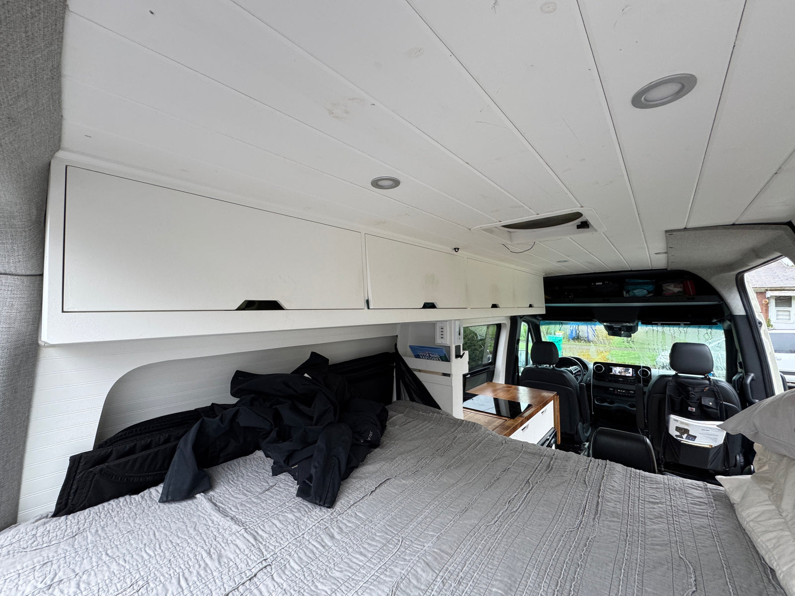 View from the back of an RV rental, showing a grey bed, white overhead cabinets, and a compact kitchen counter, leading to the front cab.