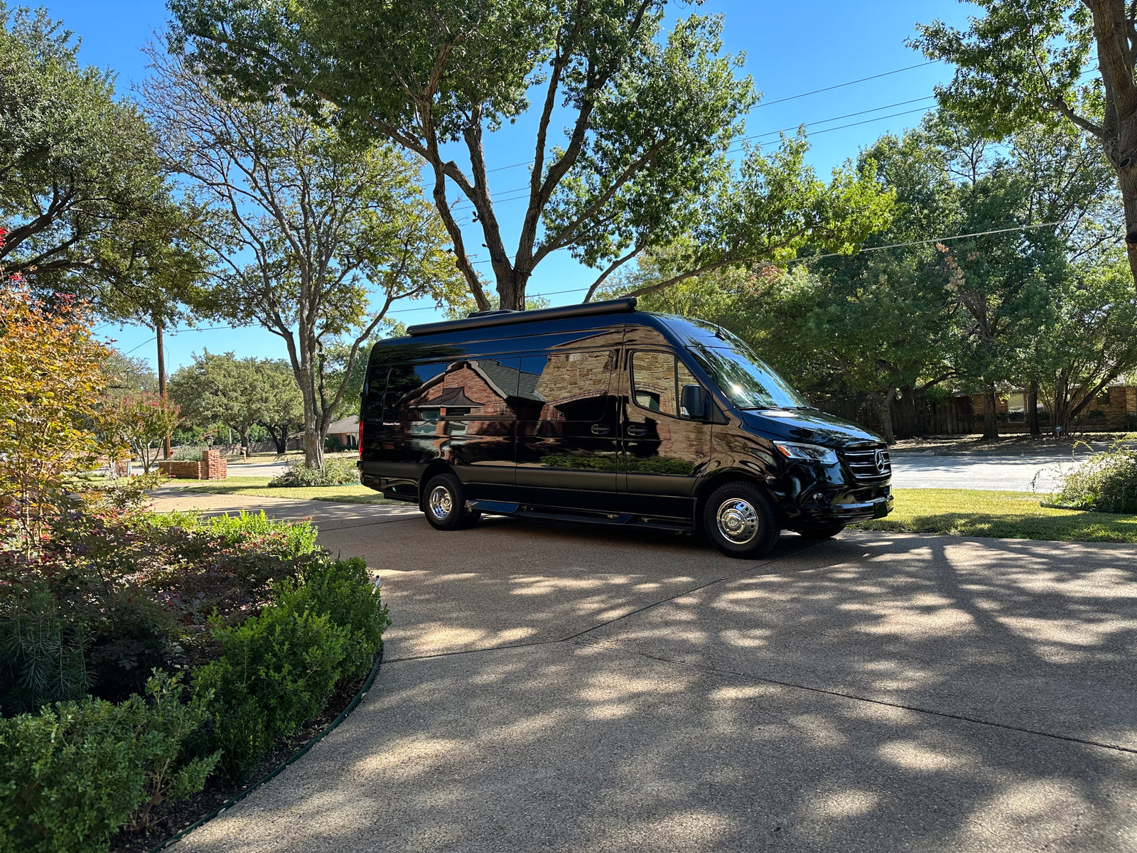 Sleek black Mercedes Sprinter van, an RV rental, parked on a sunny driveway, reflecting homes and trees on its glossy surface.