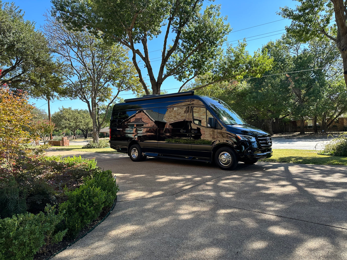 Sleek black Mercedes Sprinter van, an RV rental, parked on a sunny driveway, reflecting homes and trees on its glossy surface.