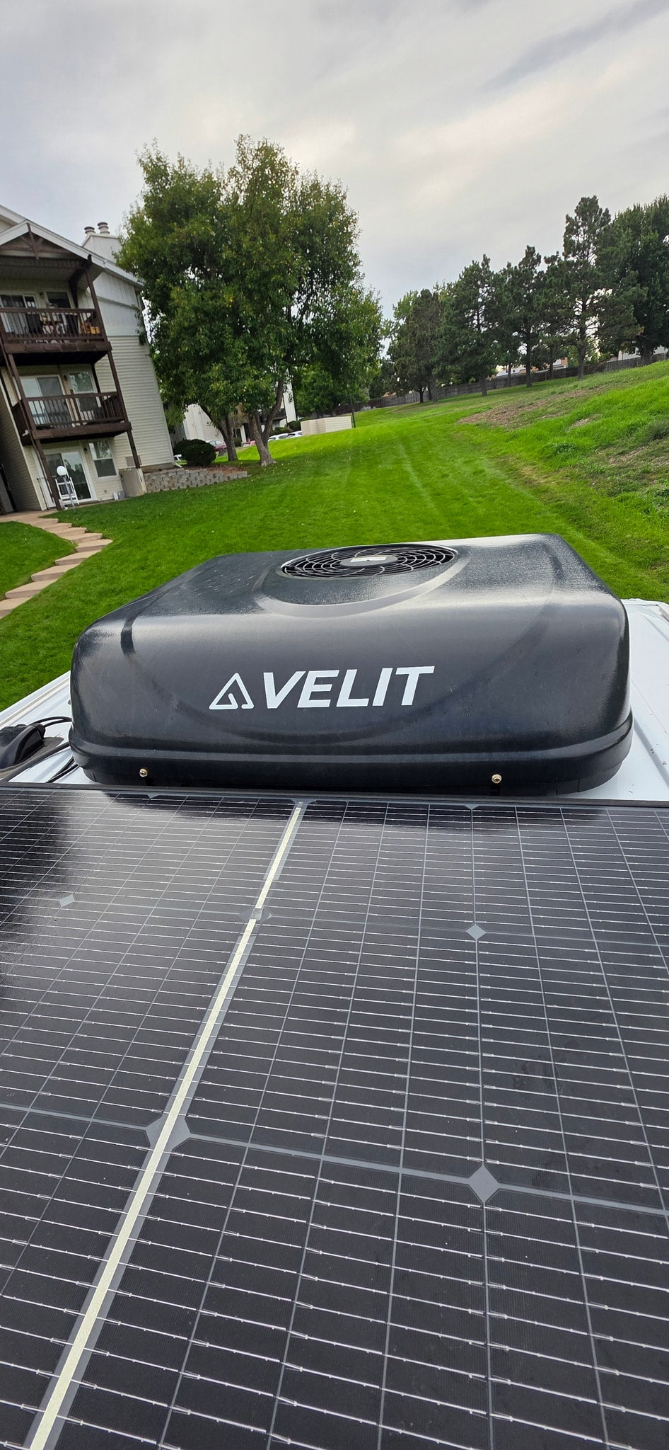 RV rental rooftop: AVELIT air conditioner and solar panels prominent. Background features an apartment building, green grass, and trees. — photo 19