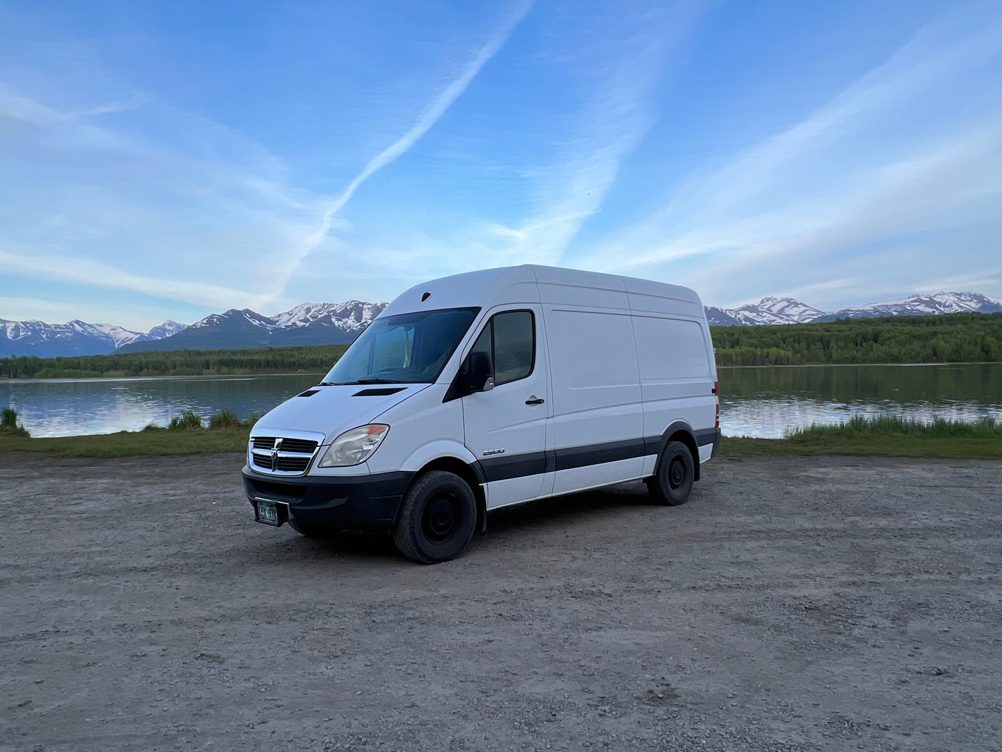 White RV rental van by a serene lake, framed by snow-capped mountains and blue sky. Your perfect adventure travel companion.