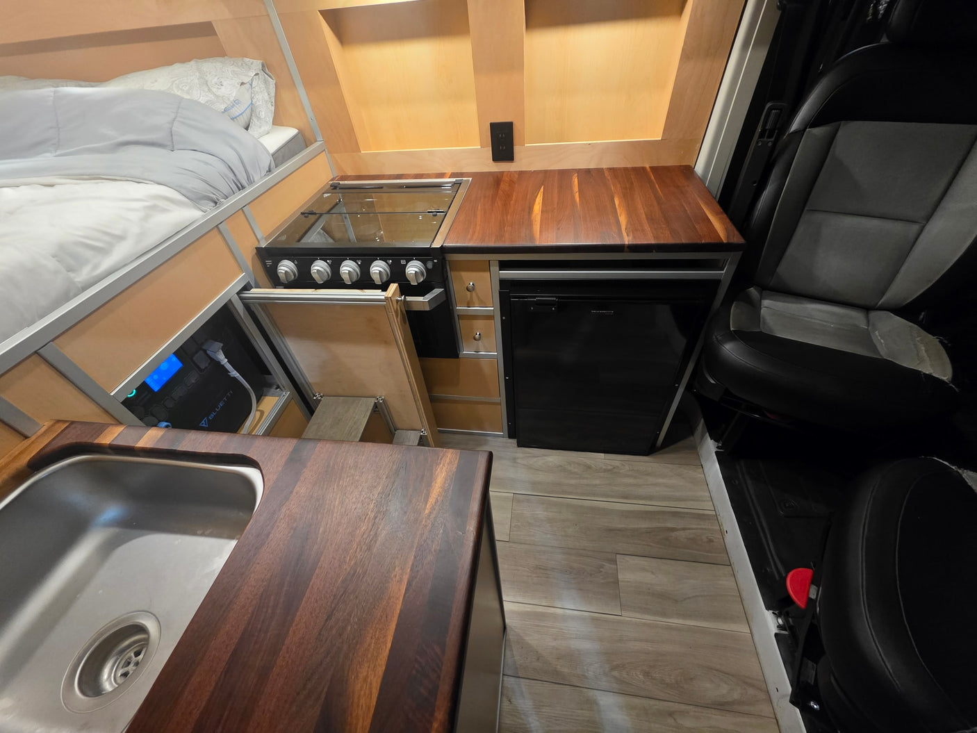 RV rental interior: cozy bed, compact kitchen with stove, sink, fridge, wooden counters, and a Bluetti power station.