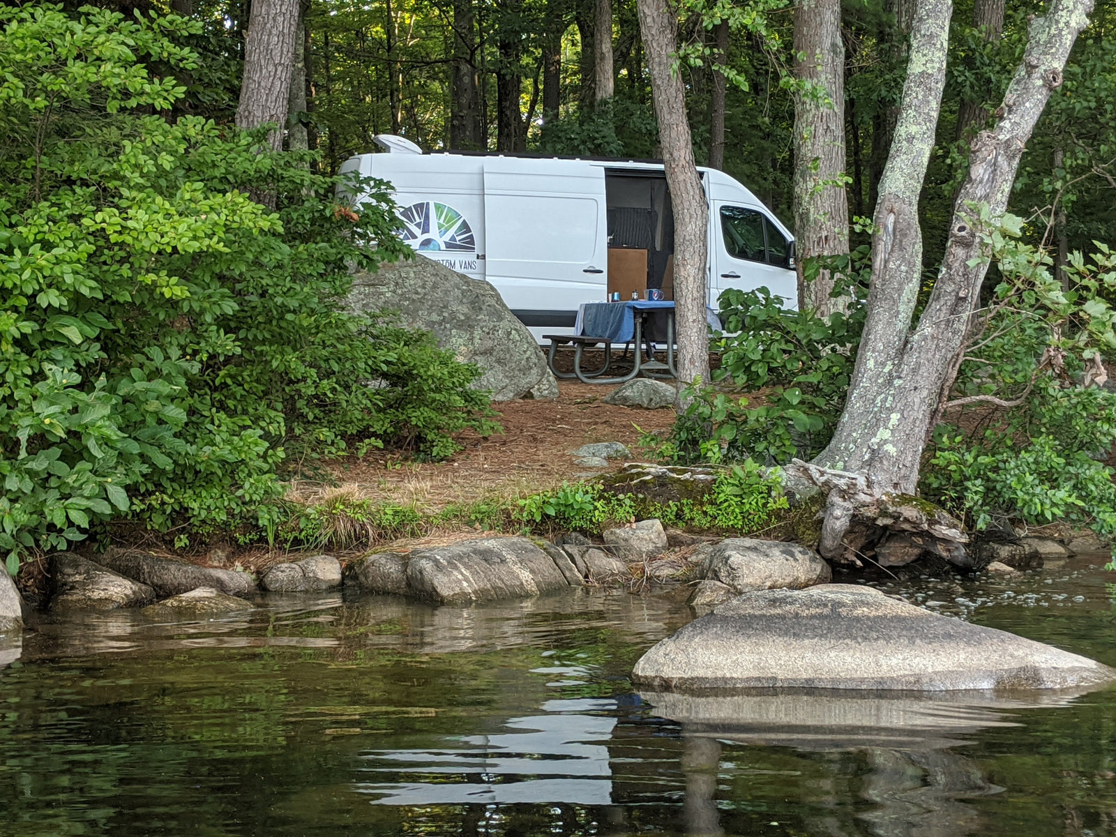 A white RV rental camper van is parked by a lake, surrounded by lush trees and rocks.