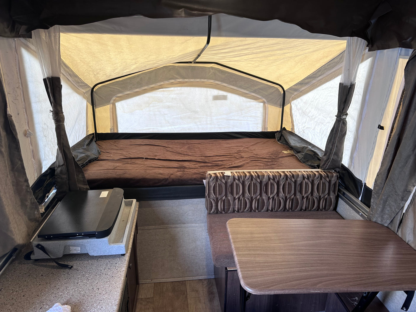 Inside an RV rental pop-up camper, showing a large bed, dining area with a table and bench, and a small kitchen counter.