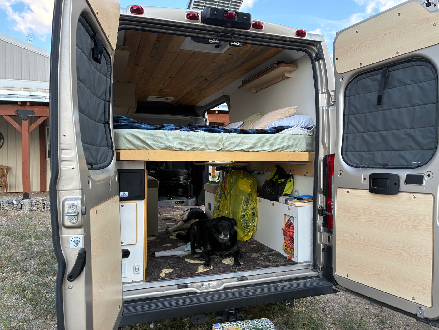 A comfortable rv rental interior, seen from the open rear doors, with a bed, storage, and a black dog relaxing inside.