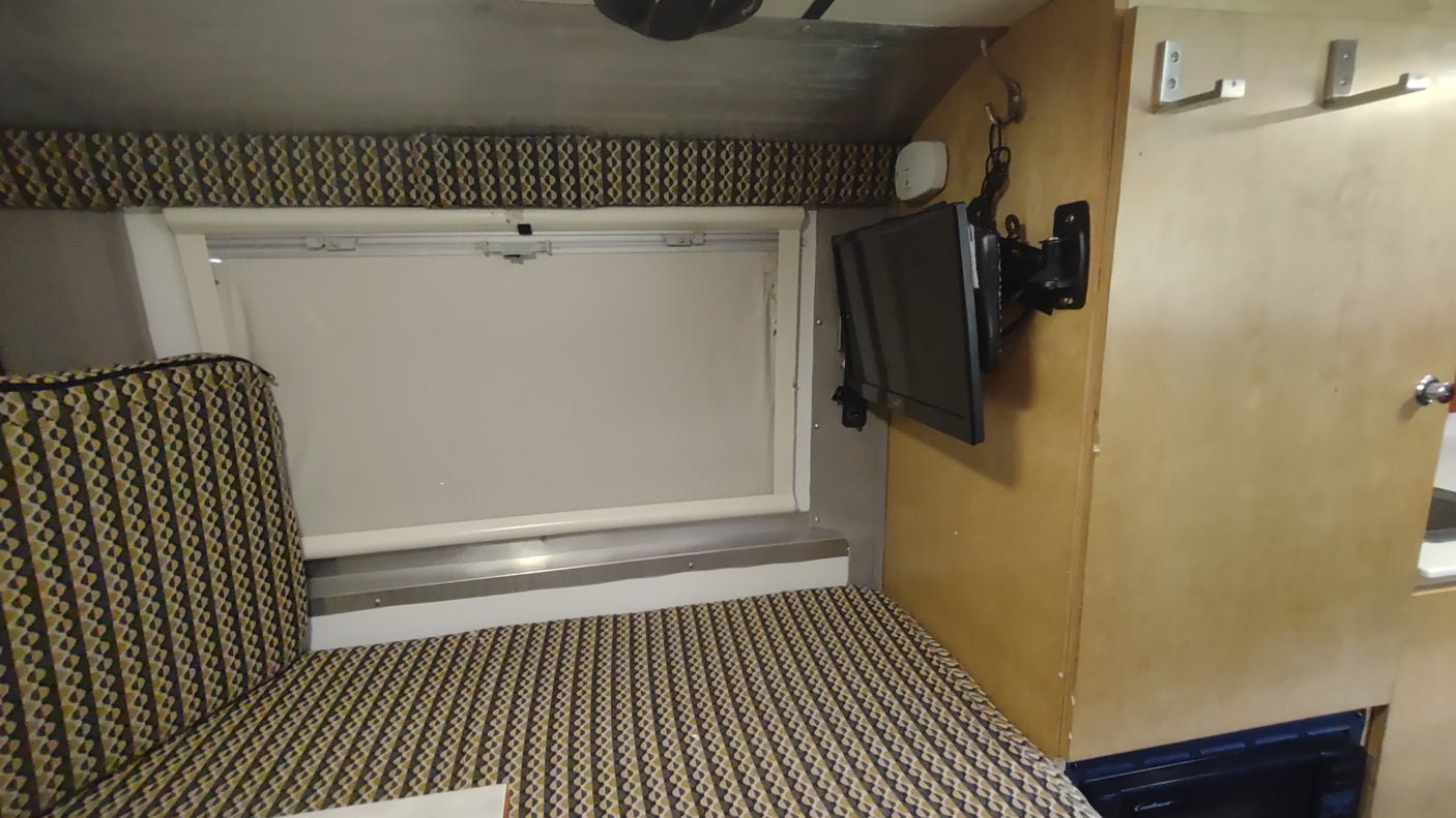 RV rental: Interior shows a cozy bed/seating with patterned fabric, a window, wall-mounted TV, and wood cabinetry.