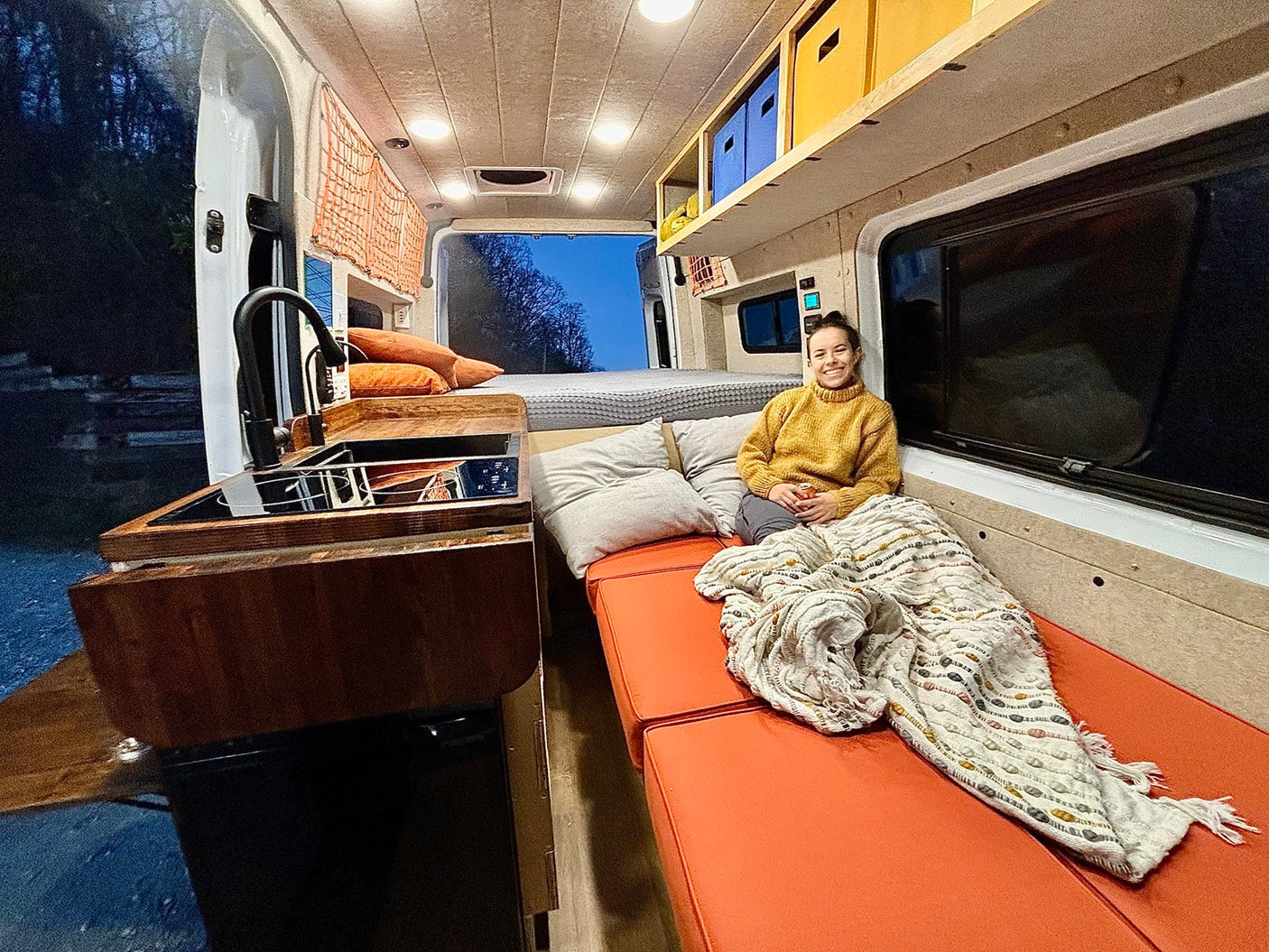 Cozy camper van interior for your next rv rental adventure. Woman smiles on bench with kitchen & bed.