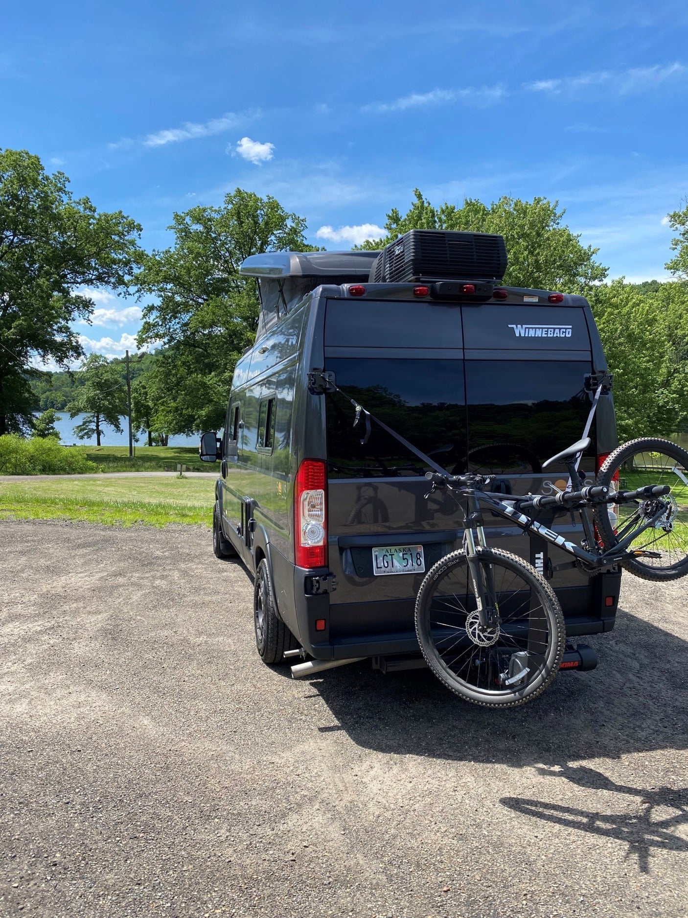 Winnebago rv rental: Dark grey camper van with a bike rack parked near a scenic lake on a sunny day.