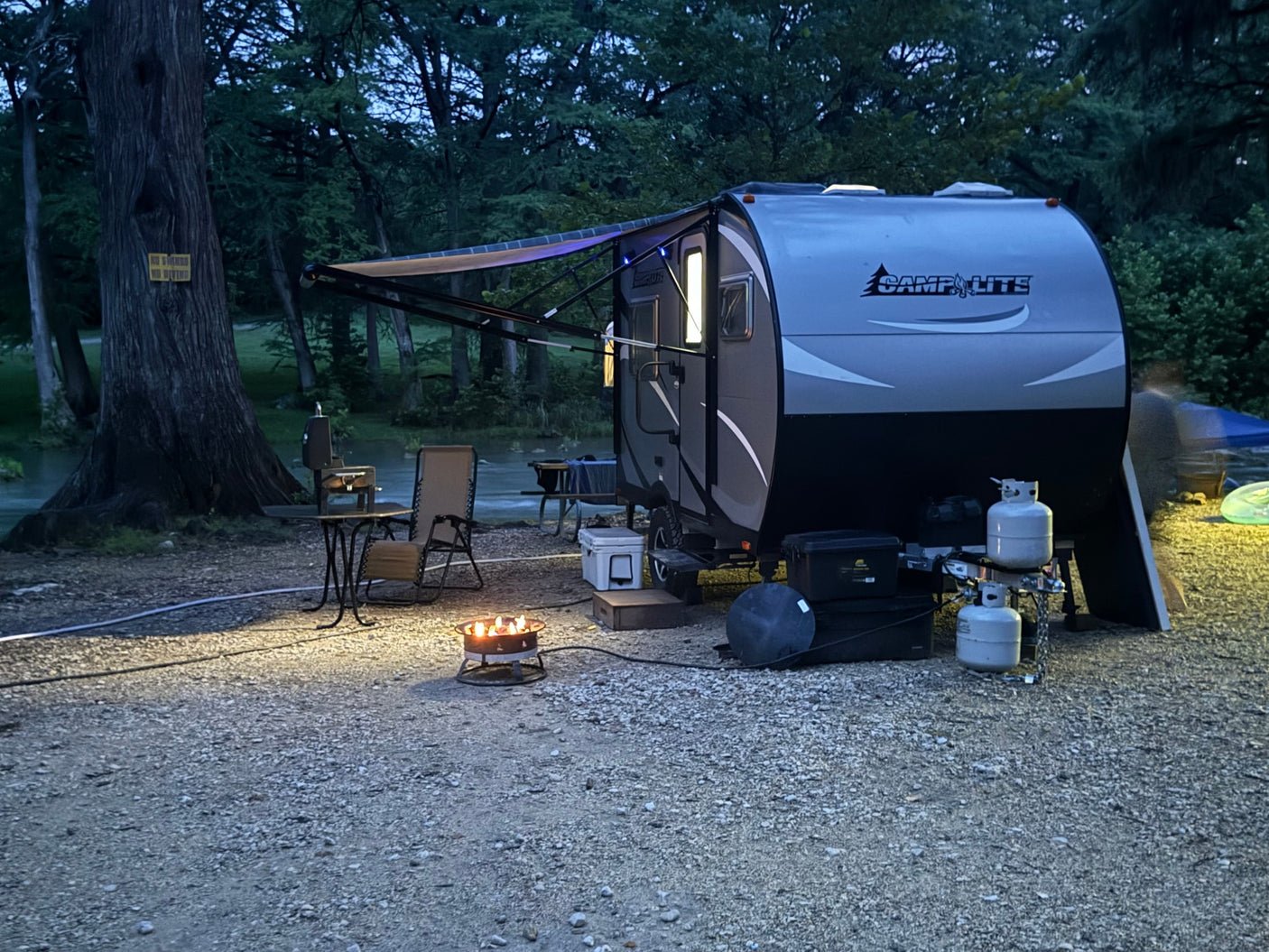 Nighttime rv rental campsite by a river with a glowing fire pit and illuminated travel trailer, perfect for a peaceful evening.