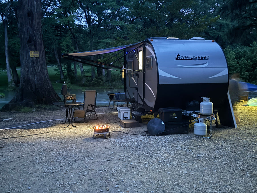Nighttime rv rental campsite by a river with a glowing fire pit and illuminated travel trailer, perfect for a peaceful evening.