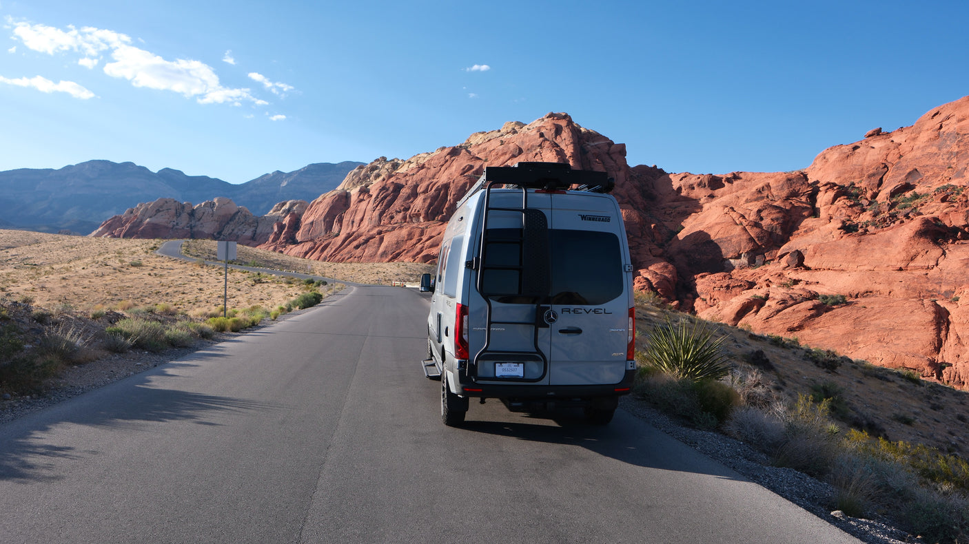 A grey Winnebago RV rental on a desert road, flanked by vibrant red rock mountains under a clear blue sky.
