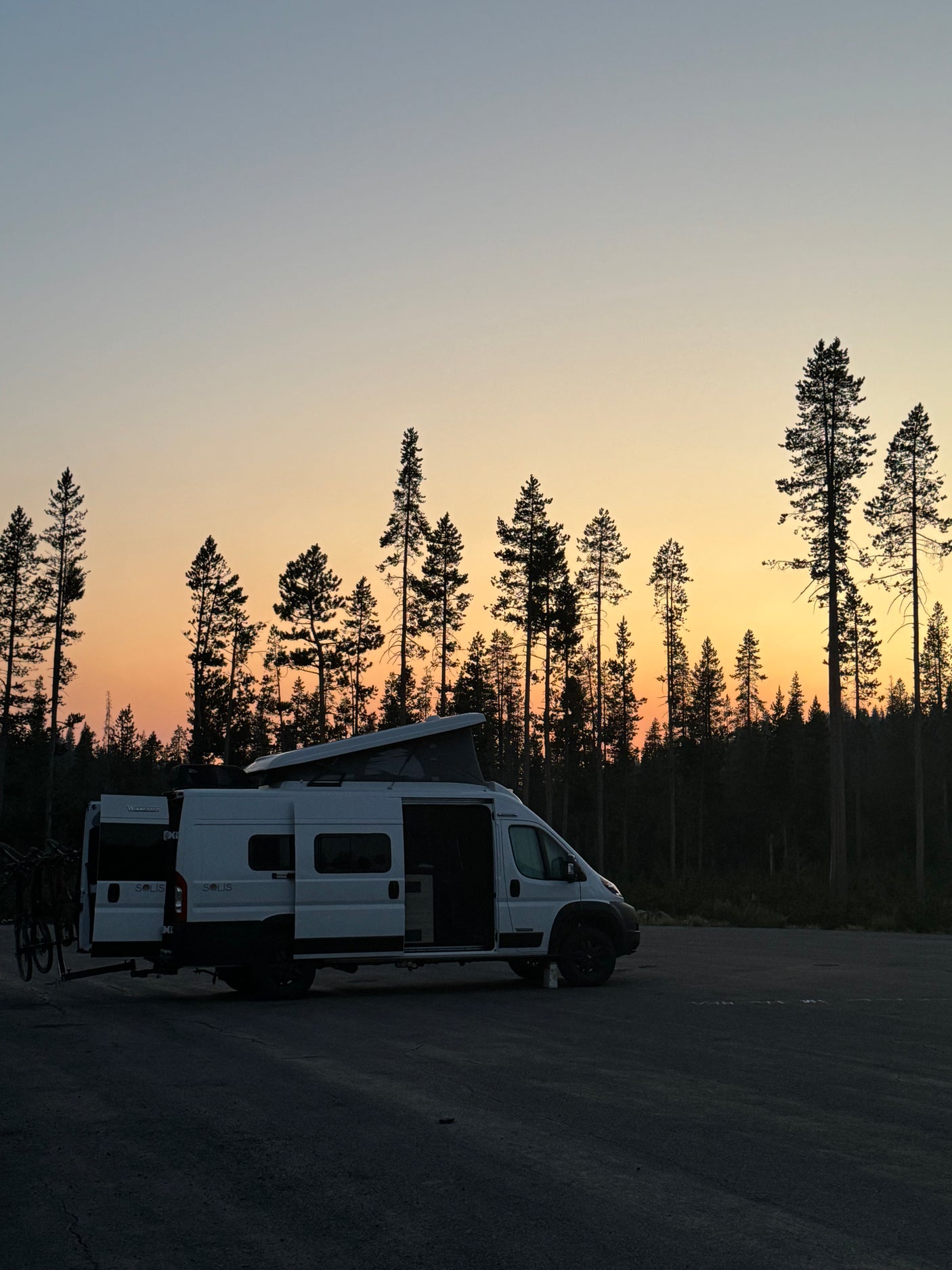 White RV rental camper van with bikes, parked in a forest at sunset with a vibrant orange and blue sky.
