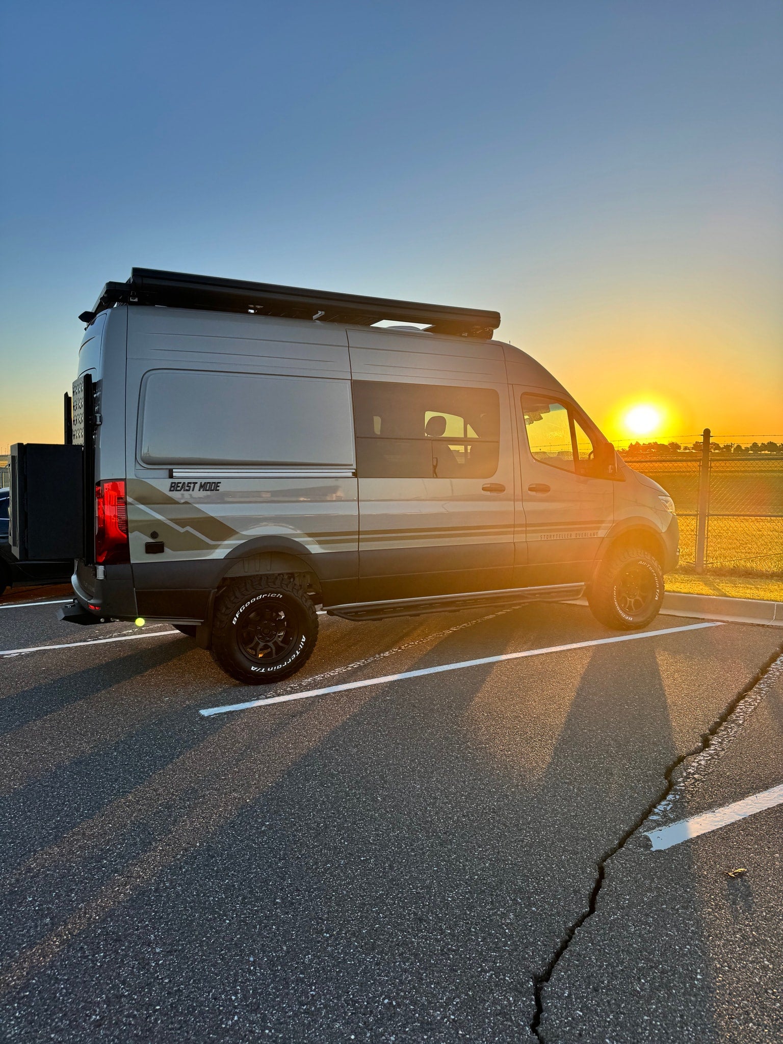 Golden hour adventure: A rugged grey Storyteller Overland RV rental van with off-road tires, parked on asphalt at sunset.