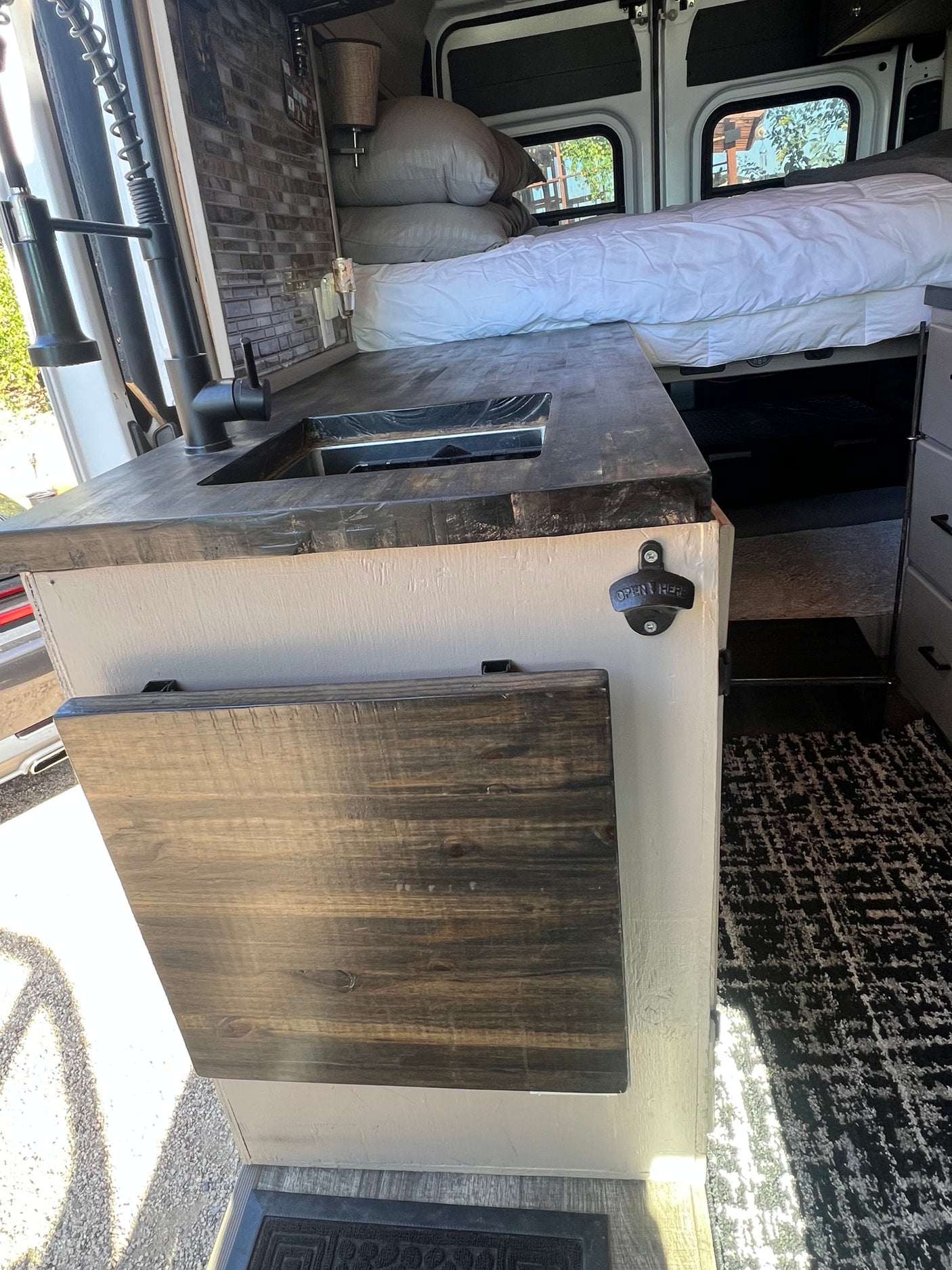Inside this RV rental: kitchenette with sink, fold-down table, bottle opener, and a cozy bed with white bedding.