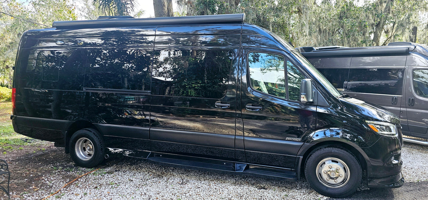 Sleek black Patriot RV rental: A Mercedes-Benz Sprinter van with an awning & side steps, parked on gravel.