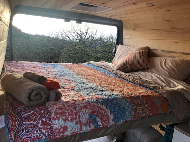 Comfortable rv rental bed with colorful quilt, rolled towels, and a beautiful natural view from the large window.