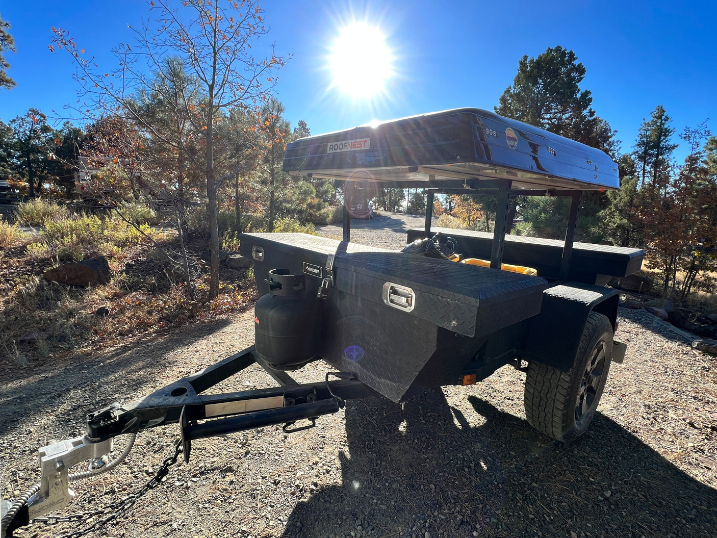 Black camping trailer with a Roofnest rooftop tent, propane tank, and toolbox on a gravel path. Great rv rental for outdoor trips.
